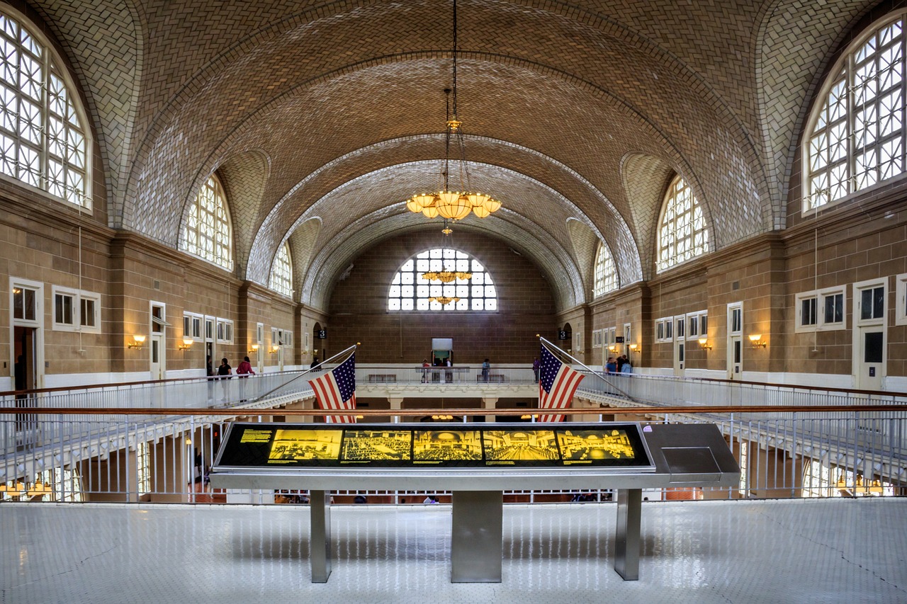 Interior of Ellis Island immigration museum with vaulted tiled ceiling, arched windows, American flags, and a digital display kiosk in the foreground.