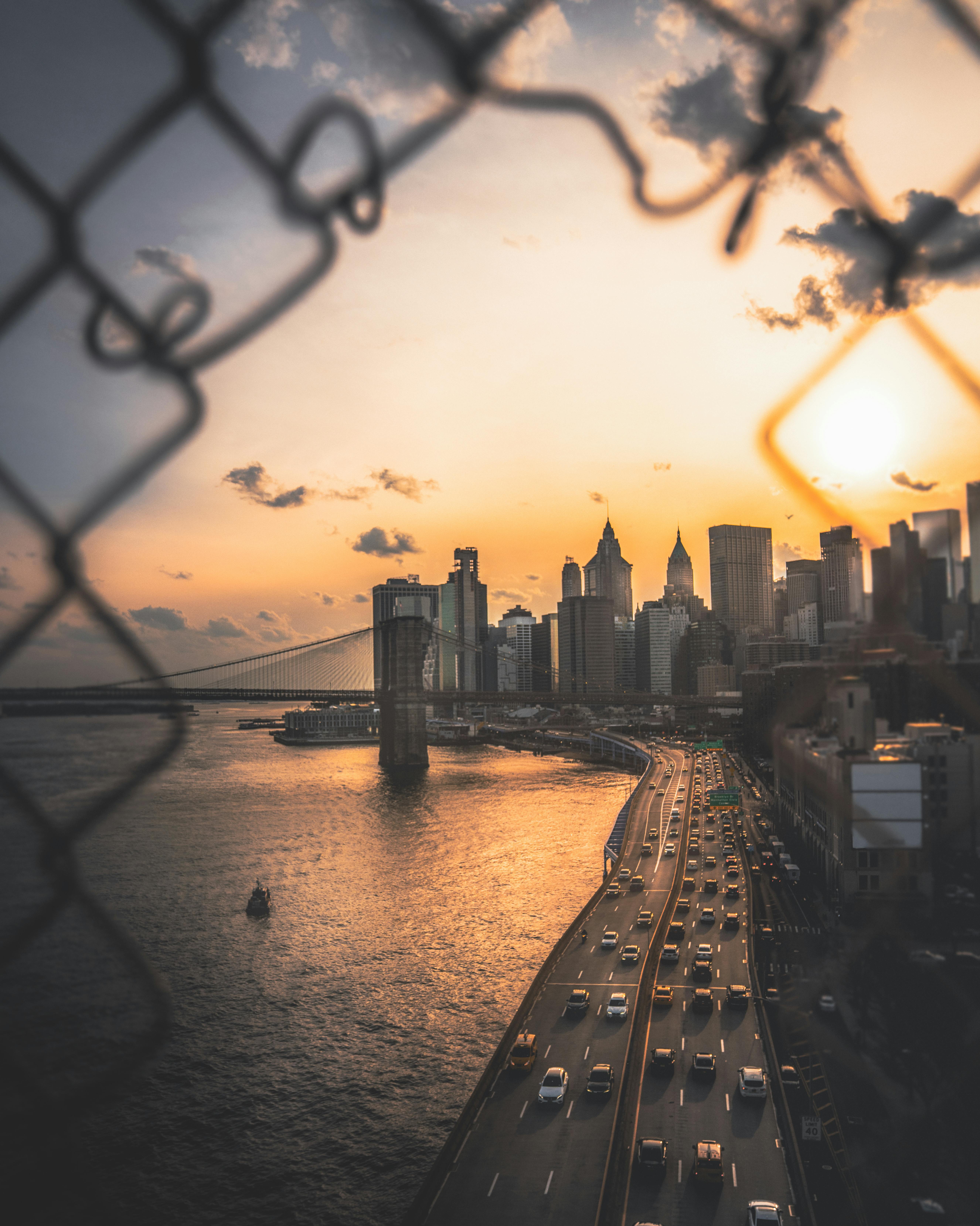 New York City skyline at sunset, viewed through chain-link fence, with Brooklyn Bridge and busy highway visible.