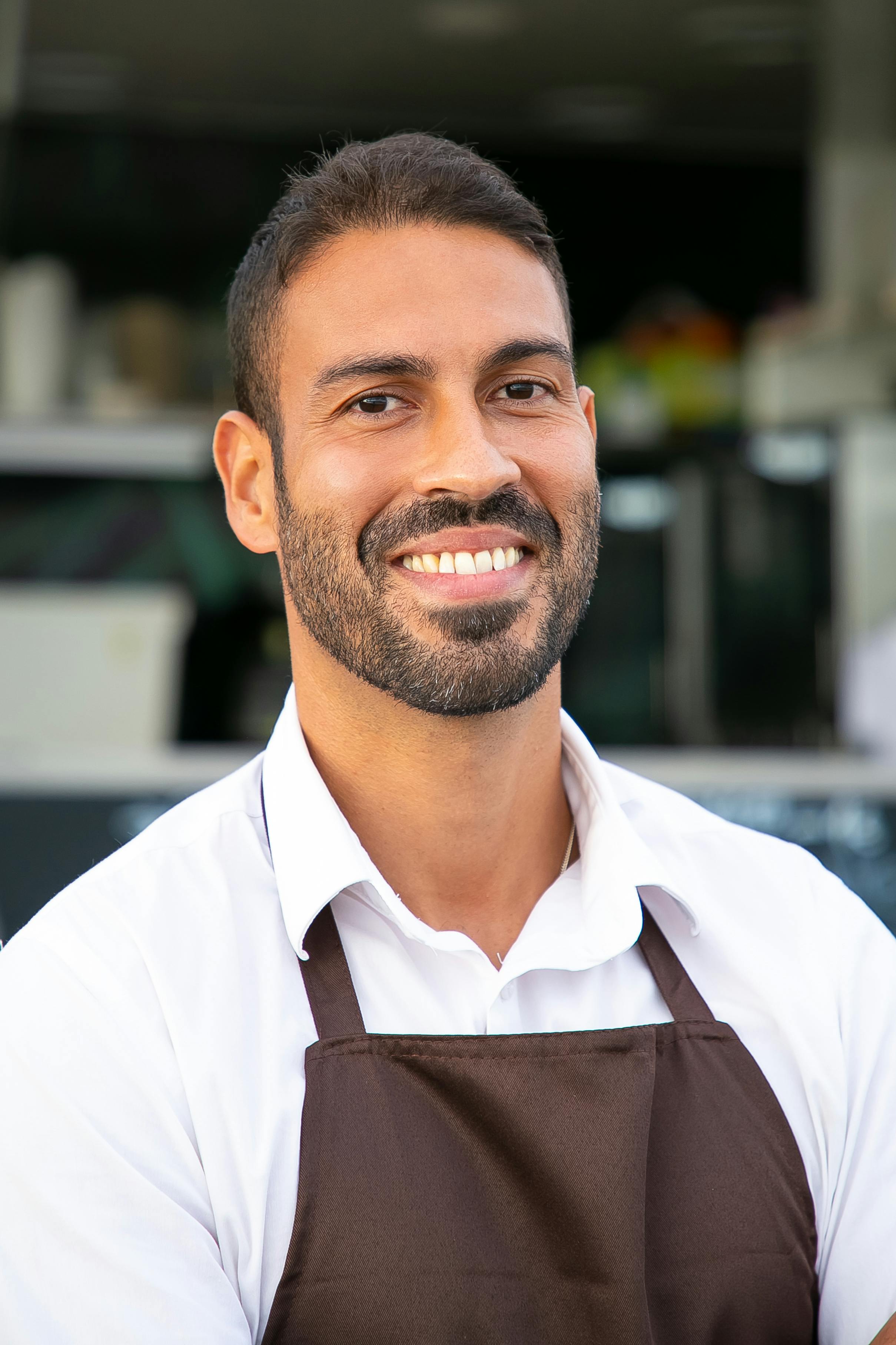 Smiling man with short dark hair and beard wearing a white shirt and brown apron, likely a restaurant or cafe worker.
