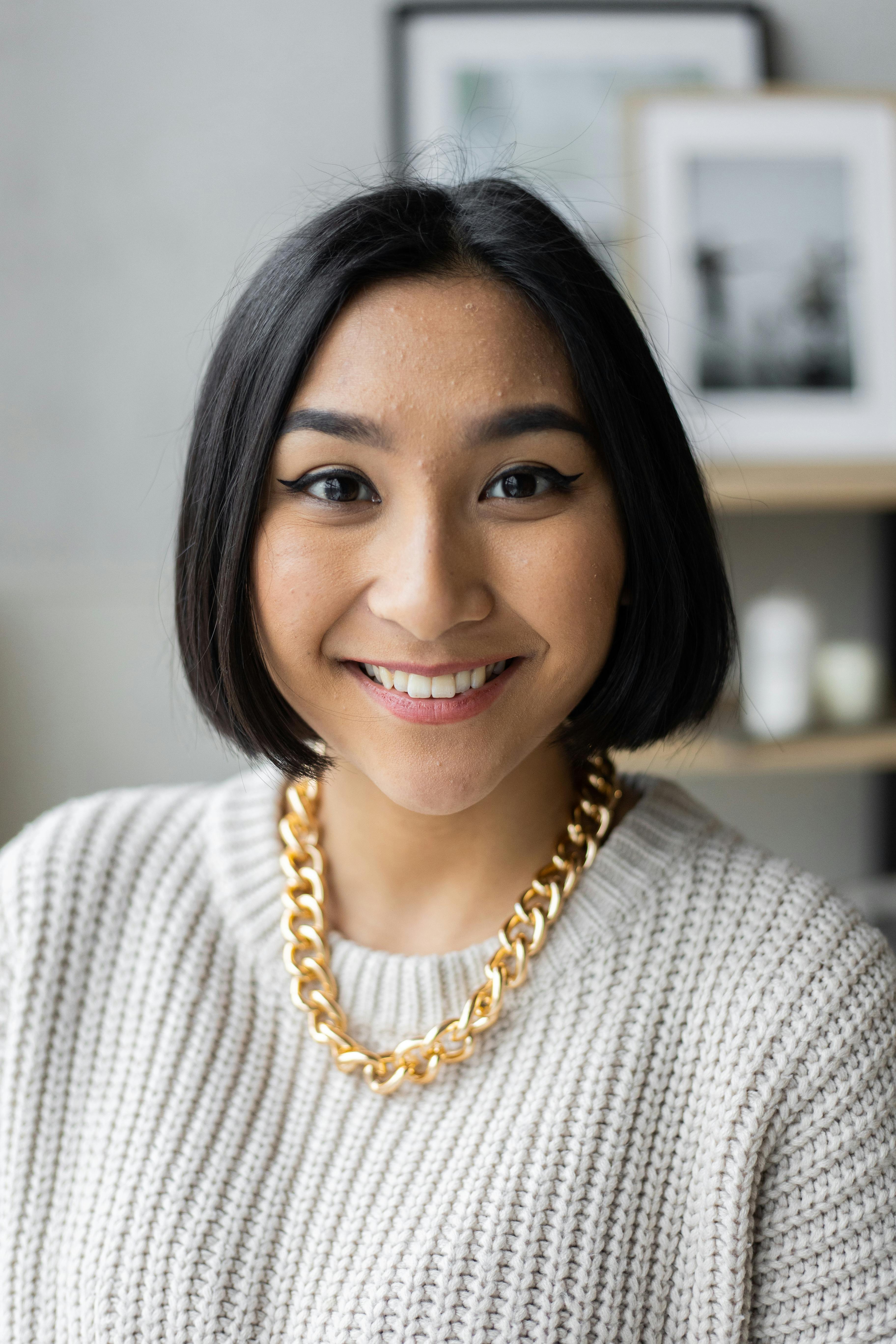 Smiling young Asian woman with short black hair wearing a white knit sweater and gold chain necklace, in a home setting with framed pictures visible in the background.